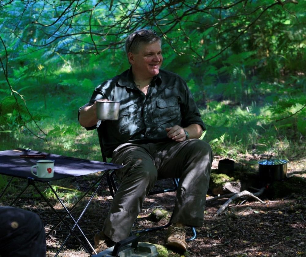 Ray Mears sitting in the woods smiling and holding a metal cup